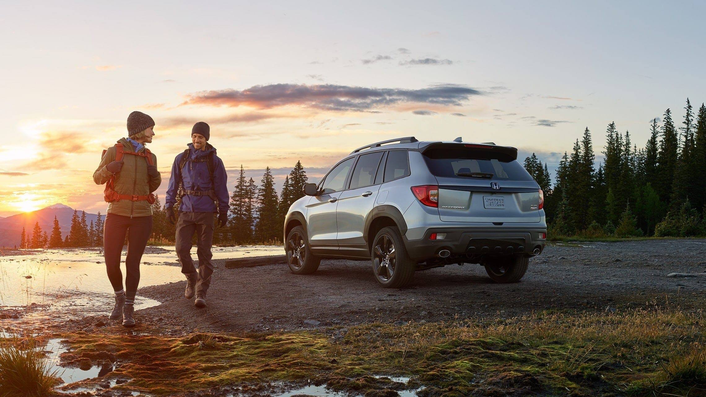 Driver-side 3/4 rear of 2019 Honda Passport Elite in Lunar Silver Metallic with accessory fender flares, at pond location with male and female hikers in foreground.