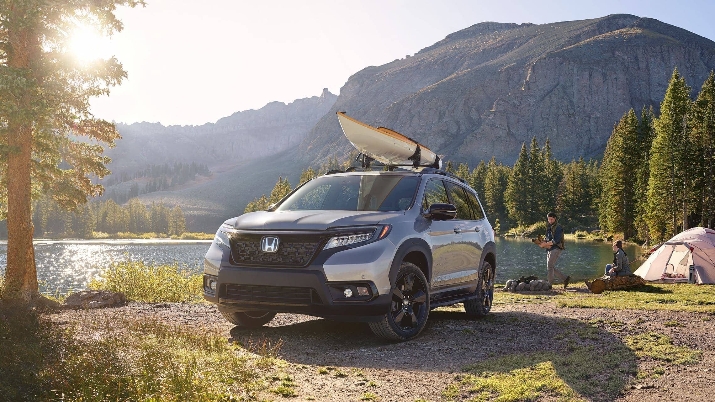 Driver-side front 3/4 view of 2019 Honda Passport Elite in Lunar Silver Metallic, with accessory kayak attachment, parked at a lakeside campground with a male and female camper.