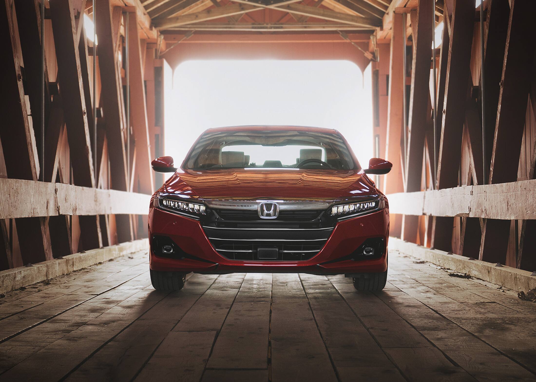 Front view of the 2022 Accord Touring 2.0T, shown in Radiant Red Metallic, parked on a wooden covered bridge with sunlight streaming in through the entrance.